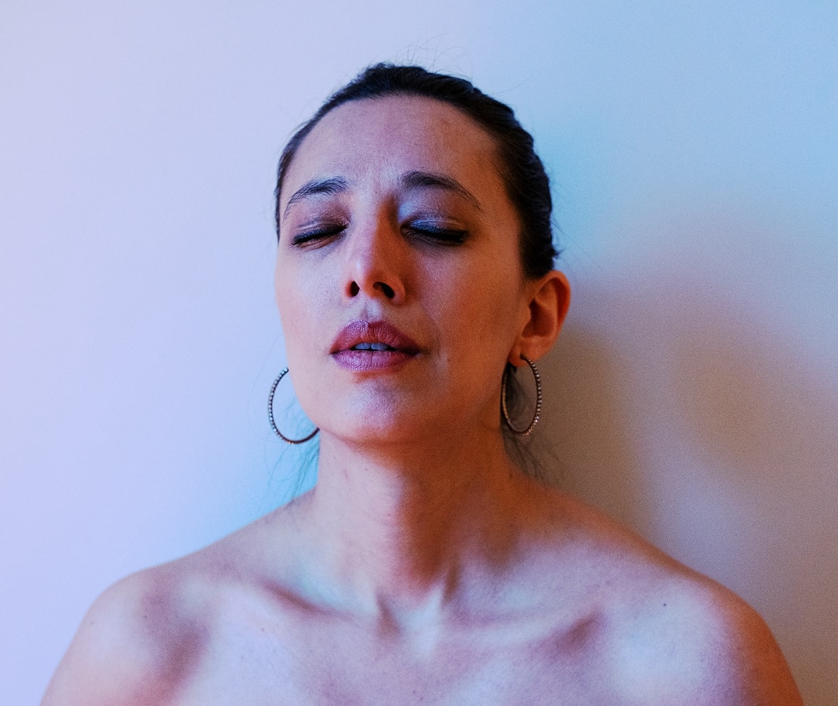 Woman with closed eyes, wearing hoop earrings and off-shoulder top, bathed in soft blue and pink light, evokes a serene expression.