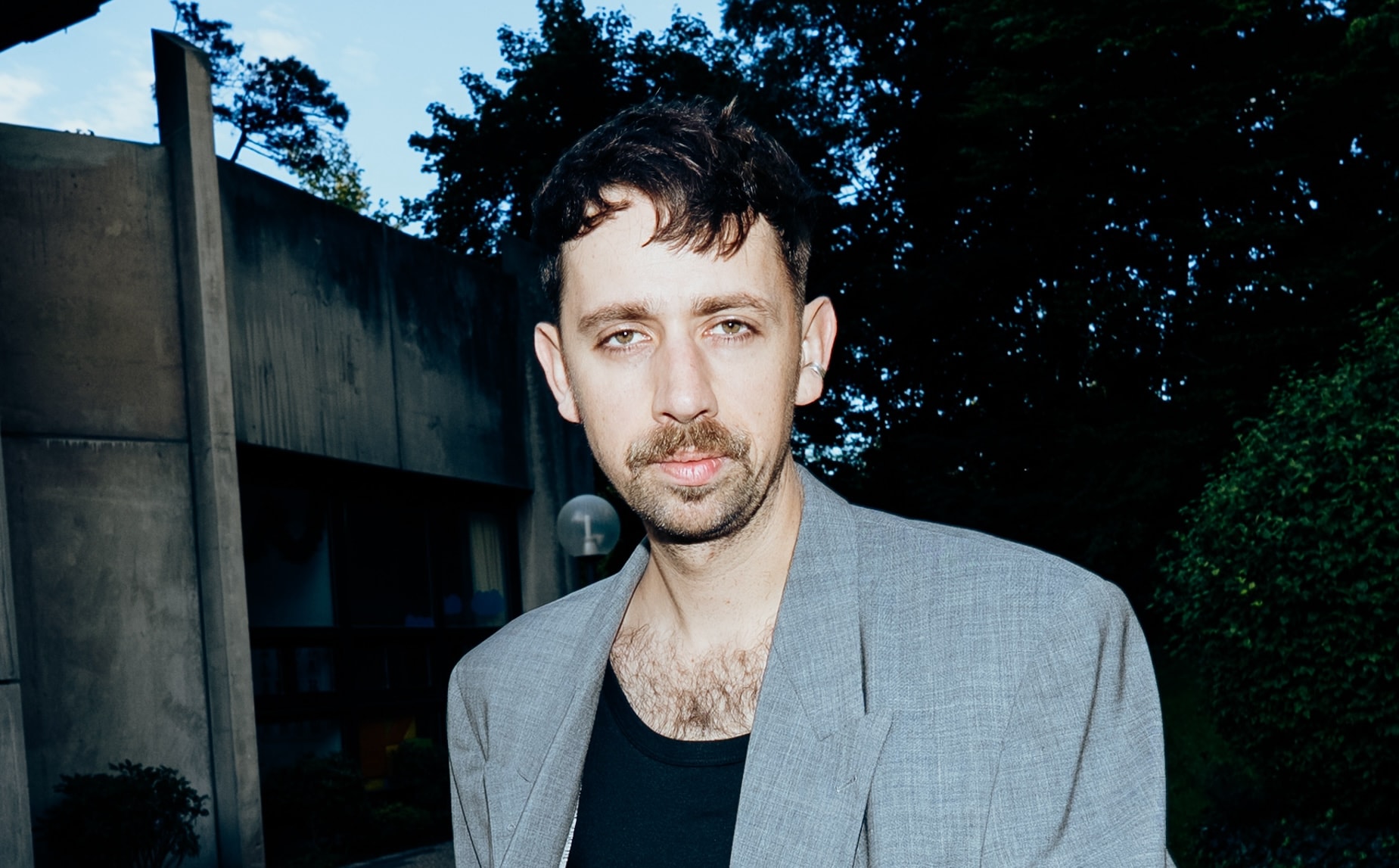 A man with short dark hair and a mustache wears a gray blazer over a black shirt, standing outdoors in front of a building and trees.