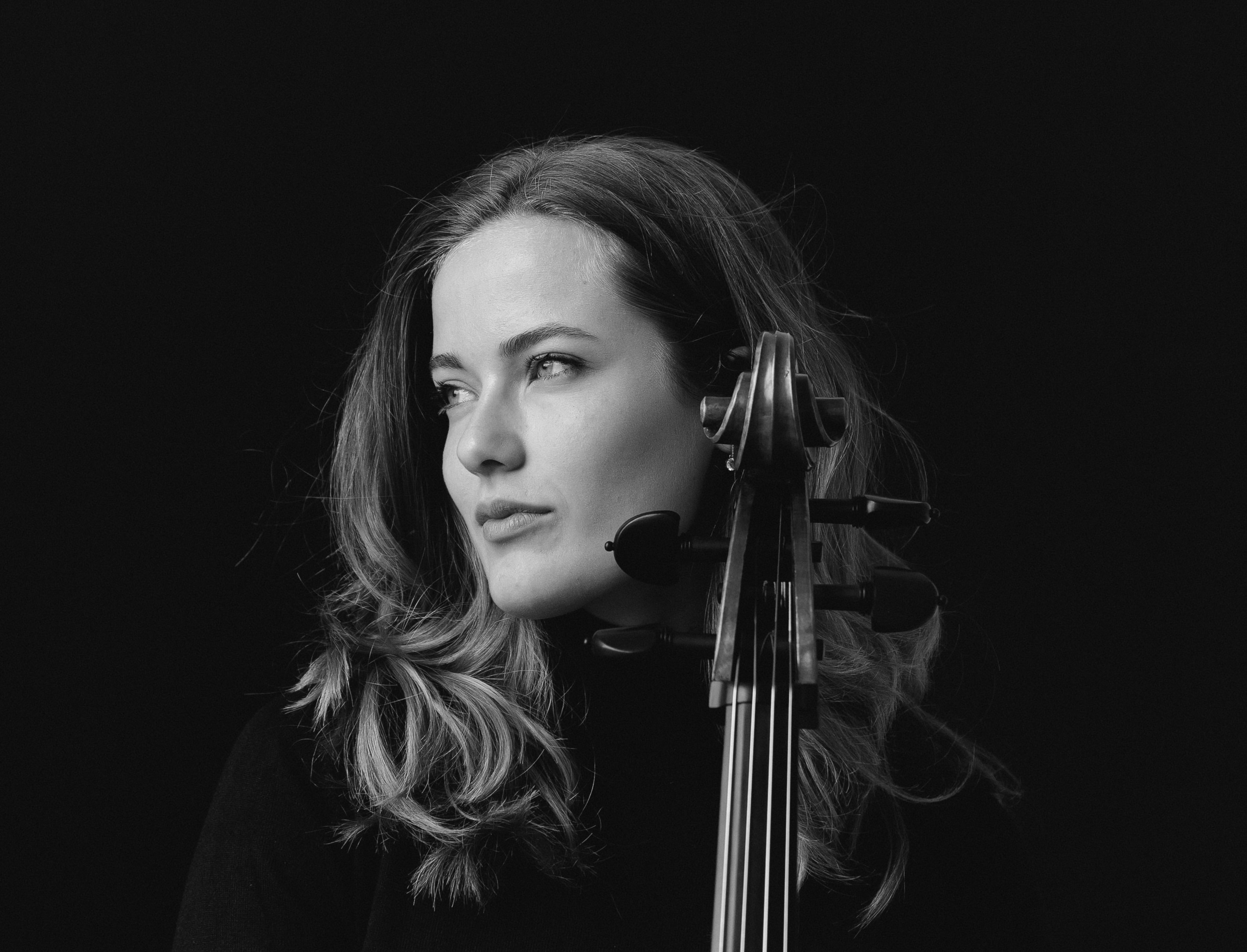 Black and white portrait of a woman with long hair, looking to the side, holding a cello against a dark background.