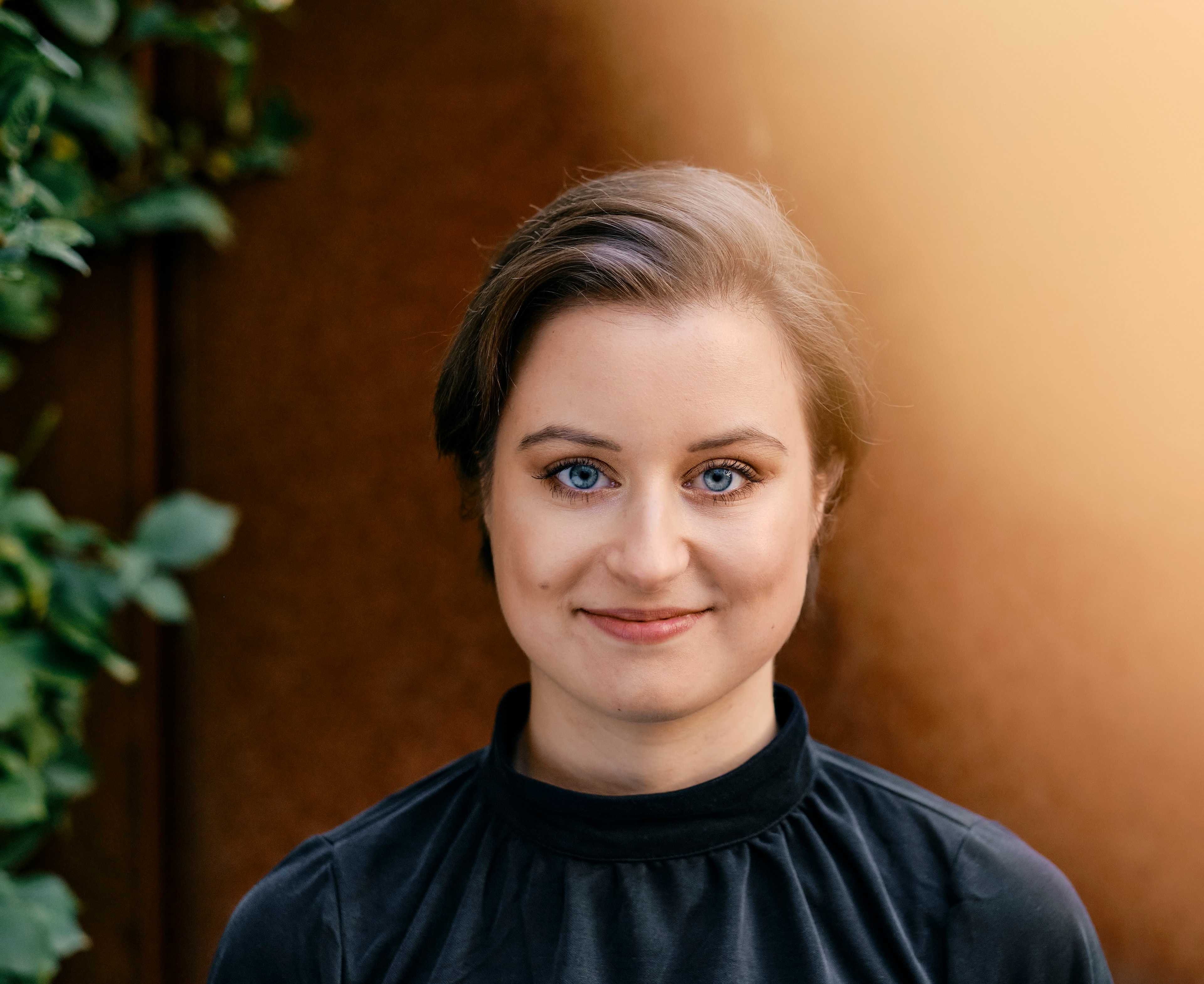 Woman with short brown hair in a black top smiling softly, with greenery on the left and a warm, blurred background.