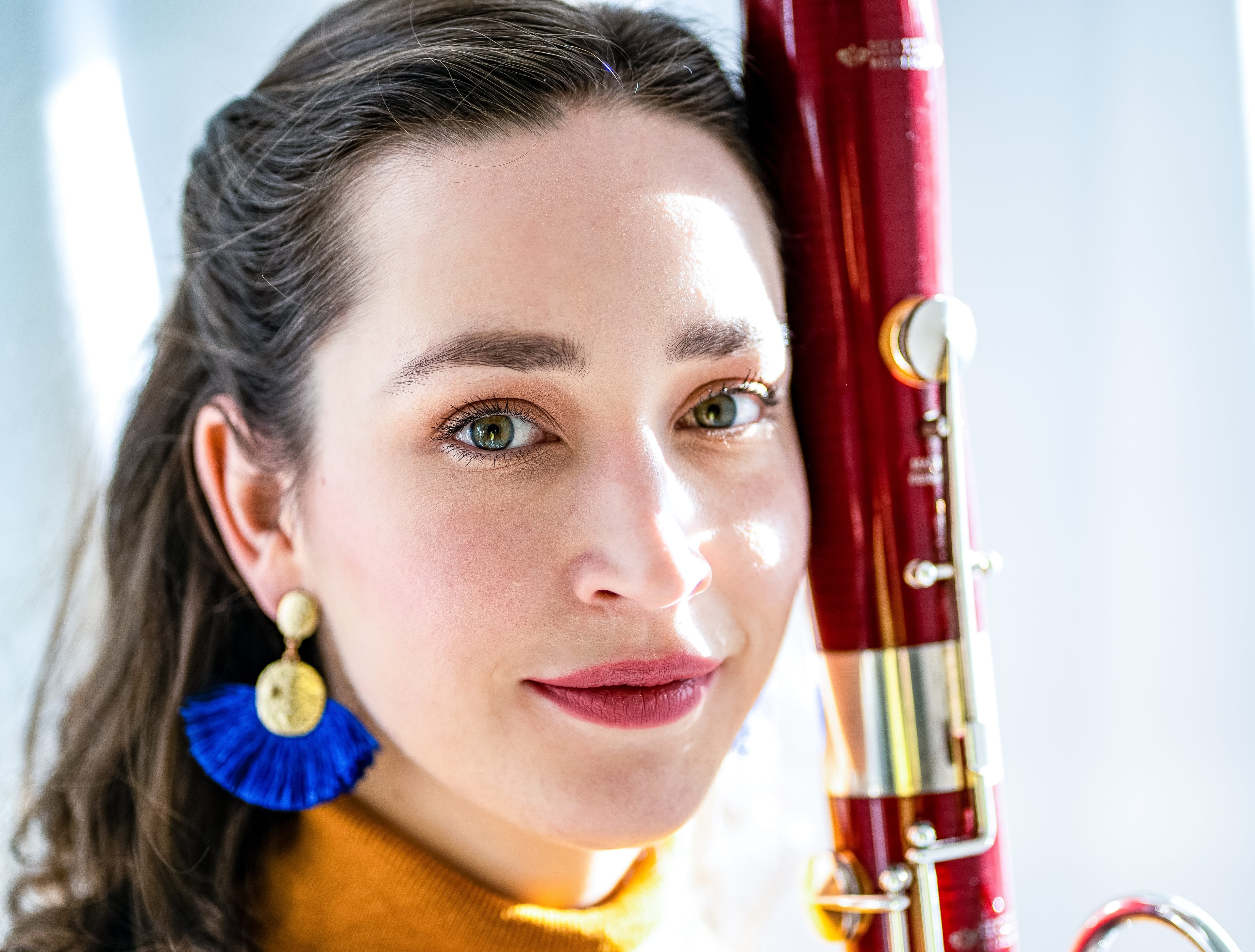 Woman with long hair and blue tassel earrings holding a red bassoon, lit by soft natural light.