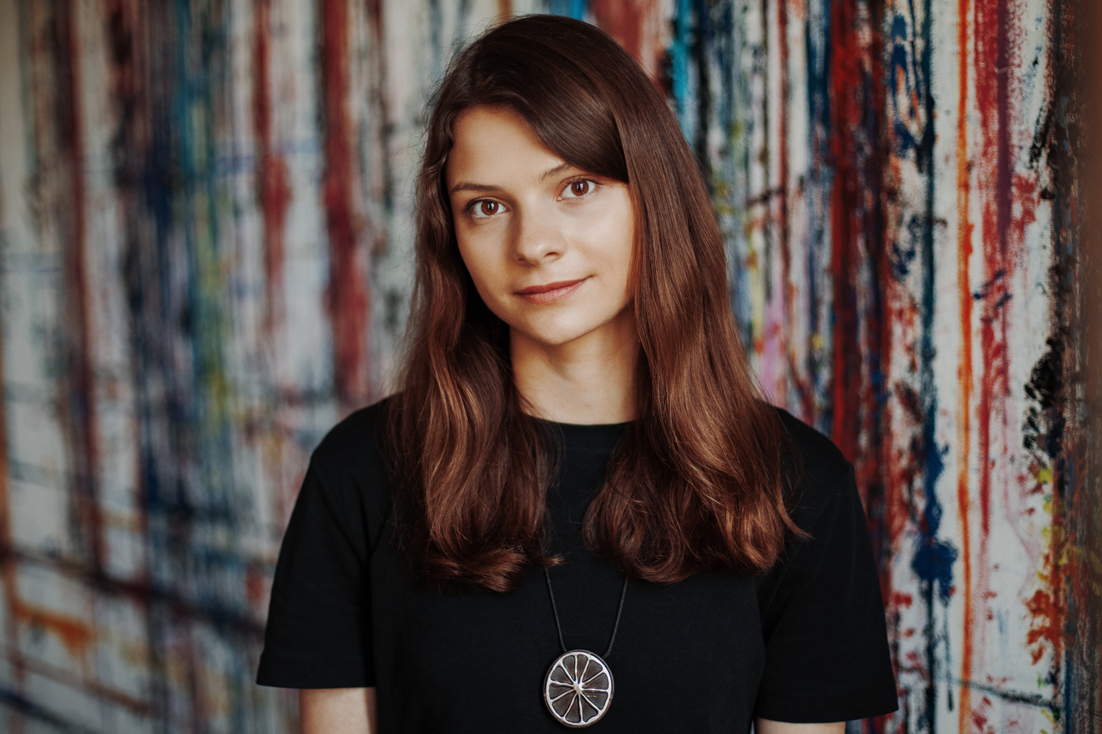 Young woman with long brown hair, wearing a black shirt and pendant, stands in front of a colorful, paint-splattered wall.