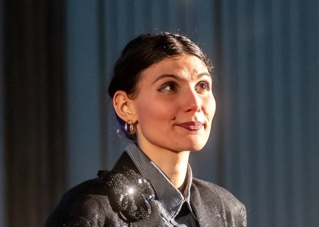 Person looking upward, wearing a dark blazer with a shiny brooch and hoop earrings, with short, slicked-back hair against a blurred background.